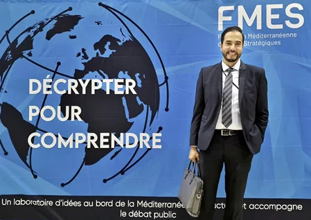 Participant en costume, mallette à la main, devant la banderole FMES « Décrypter pour comprendre »